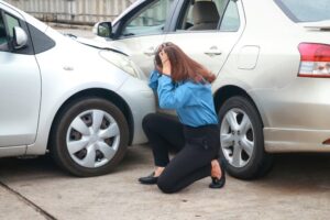 A stressed Asian woman sits on the road holding her head, looking at the crashed cars.