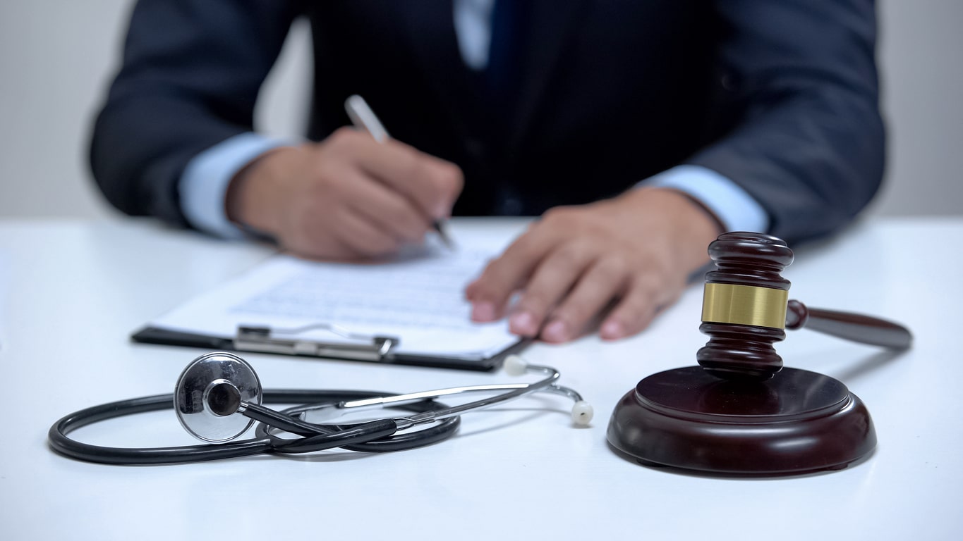 A lawyer writing on a document with a stethoscope and gavel on the table.